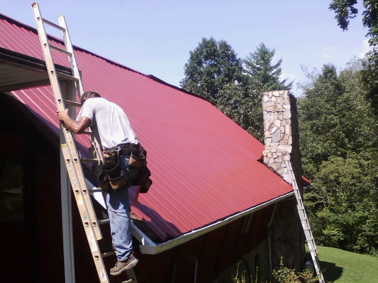 Roofer on a ladder beside a red metal roof and stone chimney — Soffit & Fascia Repair by Roofing Brothers in Yoncalla